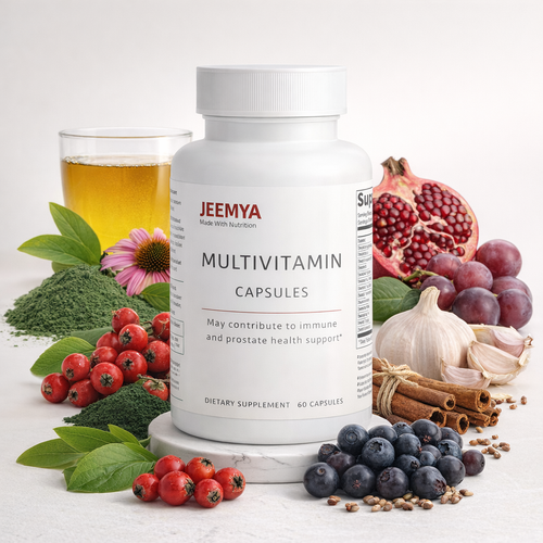 A white plastic bottle of JEEMYA Multivitamin Capsules stands on a small marble platform, surrounded by whole food ingredients against a light background. To the left is a glass of amber liquid, green powder, leaves, a pink echinacea flower, and red berries. To the right are a halved pomegranate, purple grapes, a bulb of garlic, cinnamon sticks, and a pile of blueberries in the foreground. The label reads "JEEMYA MULTIVOTAMIN CAPSULES - May contribute to immune and prostate health support.